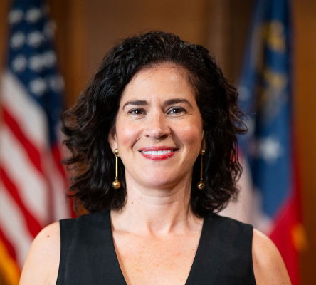 Photograph of Bárbara Rivera Holmes smiling with slightly blurred flags of United States and Georgia in background.