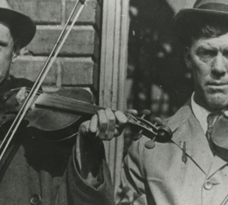 John Carson and Gid Tanner pose with their violins wearing trench coats and hats.