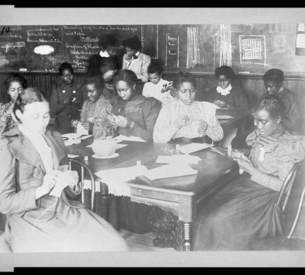A black and white photograph of a classroom of Black female students practice sewing by hand. They wear long dresses and are seated around a wood table.